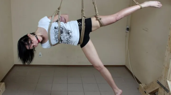 Ballgagged barefoot Ola in white t-shirt and black shorts, suspended sideways with her right leg free and pointed to the.webp