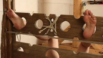 Barefoot tapegagged Irene in beige top and white shorts, lying on the floor with her feet locked in pillory, is getting.webp