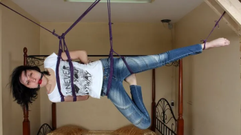 Ballgagged barefoot Ola in white t-shirt and blue jeans, suspended with violet hemp rope sideways, is tickled by camera.webp