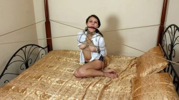 Ballgagged topless collared Roxana in white unbuttoned shirt and sport shorts, tied sitting on the bed with her feet cro.webp