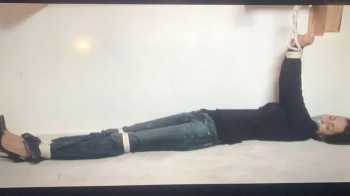 Tape-gagged brunette Gia Mancini lay stretched out and writhing on the floor with her arms tied overhead to a wooden bar.webp